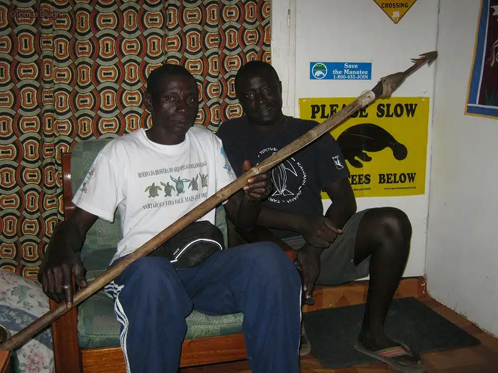 Two men sitting in a room with Save the Manatee Club materials in the background, the person on the left is holding a harpoon.